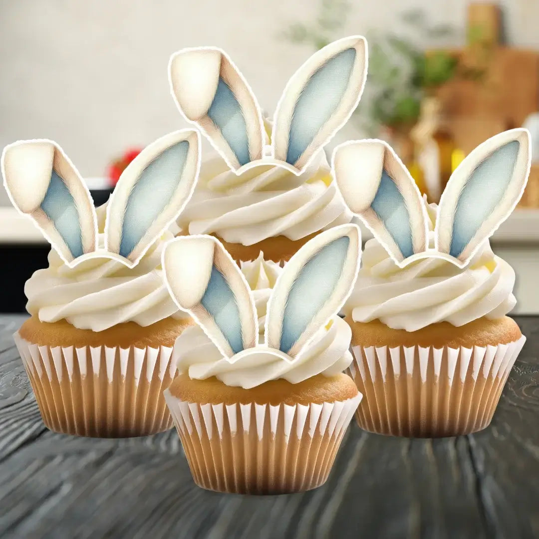 Blue and white wafer bunny ears, placed on hite frosted cupcakes, on a kitchen counter.