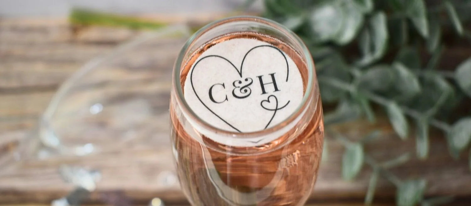 Black heart with initials printed on a wafer drinks topper, placed in a glass of rose champagne on a decorated table.