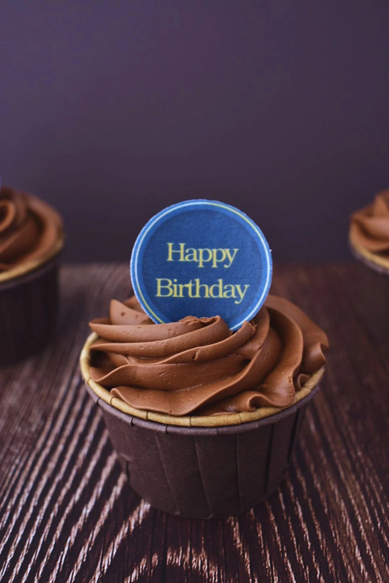 Blue cupcake topper with gold 'happy birthday font, placed on top of a chocolate frosted cupcake.