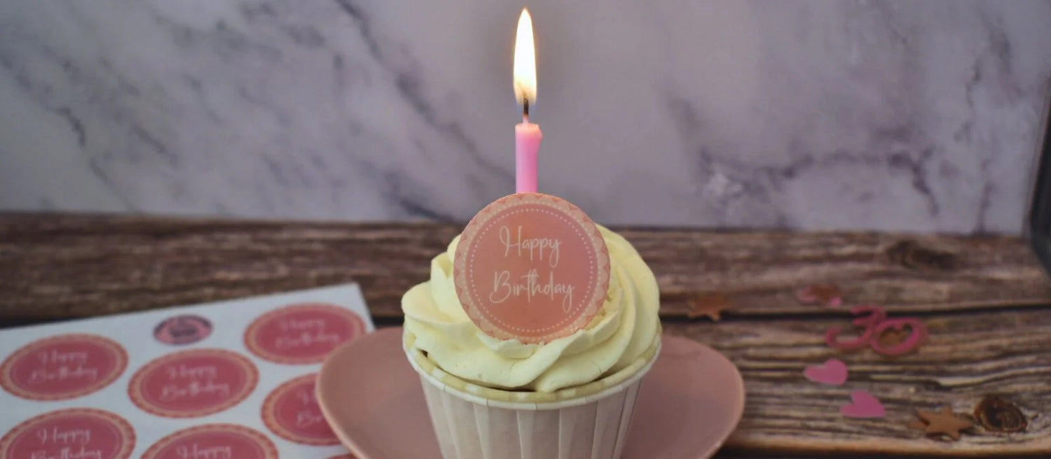 White frosted cupcake with a pink 'happy birthday' cupcake topper and a pink candle, on a pink plate and with decorations laid around.