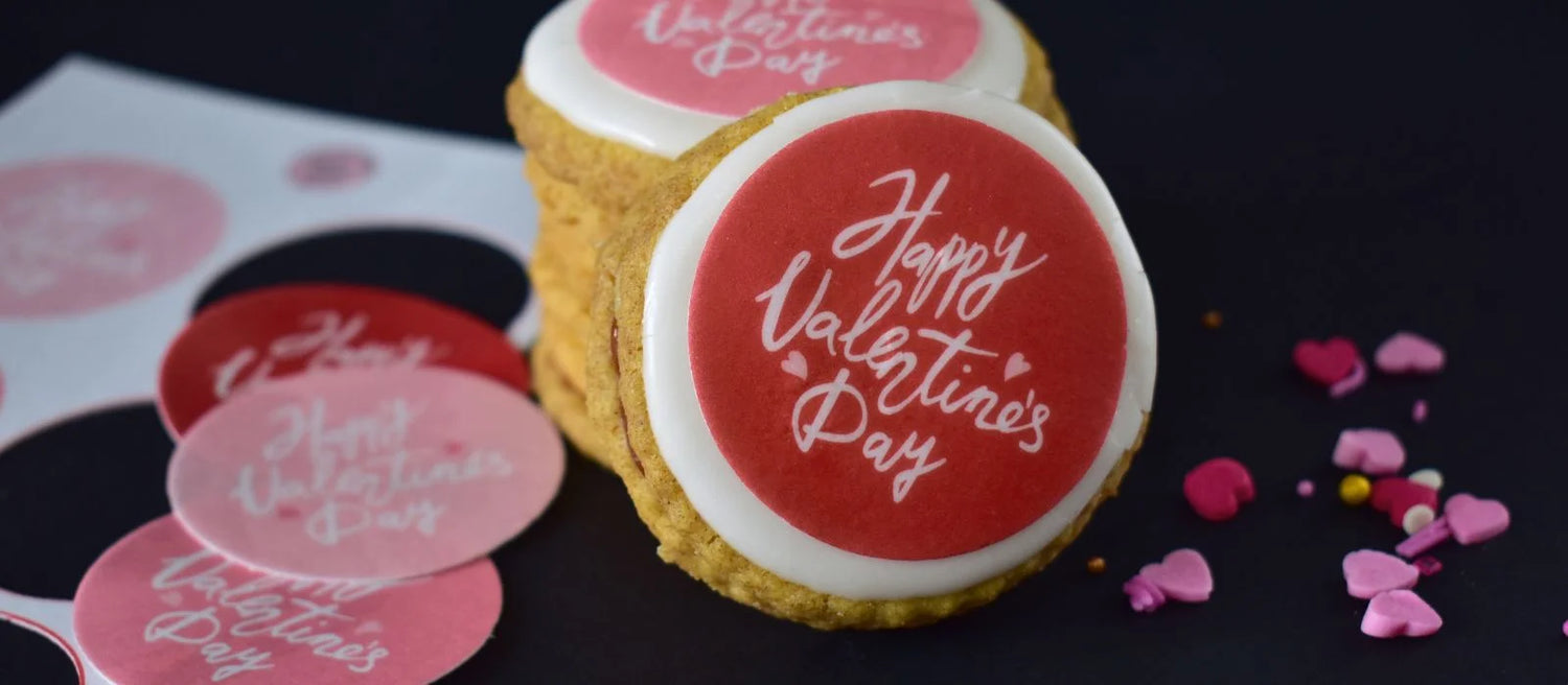 Red Valentines day edible toppers with white looping 'Happy Valentines Day' text placed on empire biscuits. There is a stack of biscuits with sprinkles and extra toppers around them.