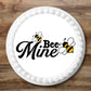 Round white cake with 'Bee Mine' text and bee graphics on a wooden surface