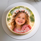 A round custom cake topper or printed cake top featuring a smiling young girl in a flower crown with the text 'Happy Birthday Sophie' on a white cake, photographed from above on a white plate.