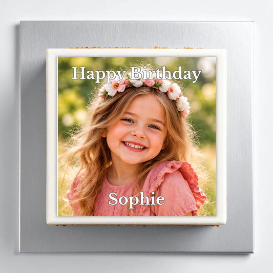 A square personalized cake or plaque with a printed photo of a smiling girl in a flower crown, bordered in white and set on a brushed silver square base. The text 'Happy Birthday' appears at the top and 'Sophie' at the bottom.