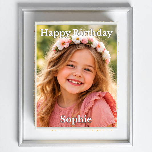 A personalized framed photo print featuring a smiling young girl with a flower crown and the text 'Happy Birthday Sophie'. The print is enclosed in a white mat and a silver frame, shown against a clean white background.