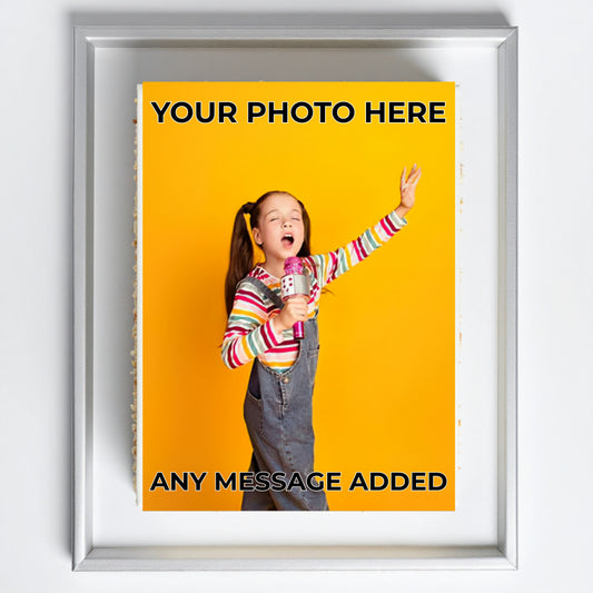 A framed customizable photo print mockup on a white wall. The print features a young girl in a colorful striped shirt and denim overalls holding a microphone against a bright yellow background, with placeholder text reading 'YOUR PHOTO HERE' and 'ANY MESSAGE ADDED'.